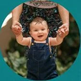A smiling baby taking steps while holding a parent’s hands
